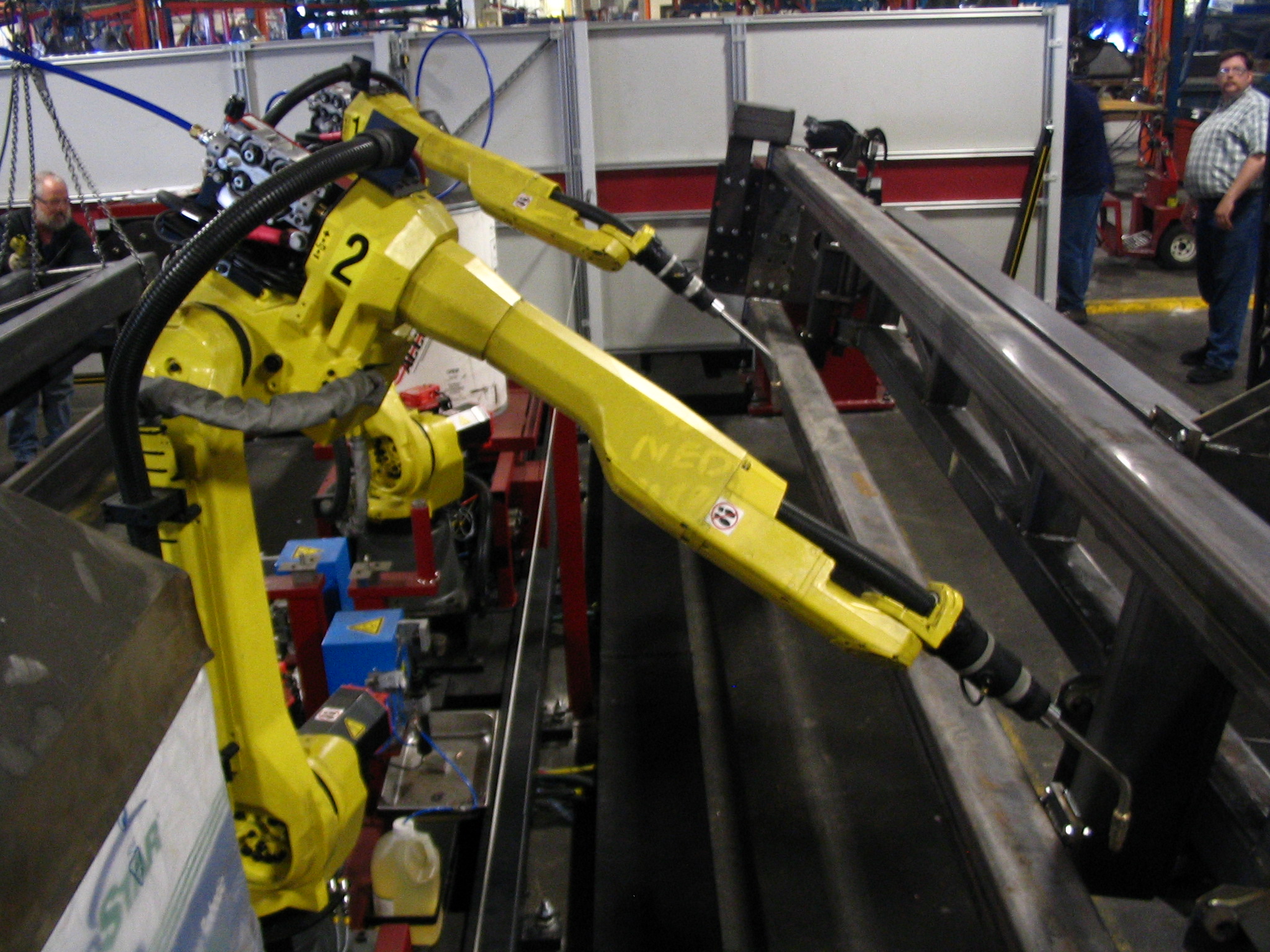 Two FANUC industrial welding robots positioned to weld a metal part inside a factory cell