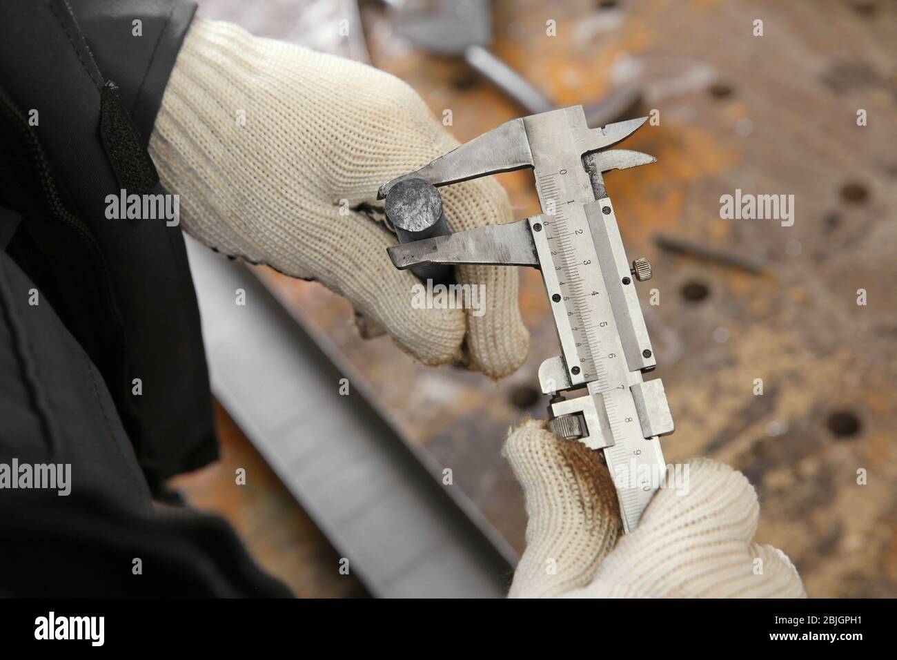 Gloved worker measuring a steel bar with a vernier caliper in an industrial metalworking shop